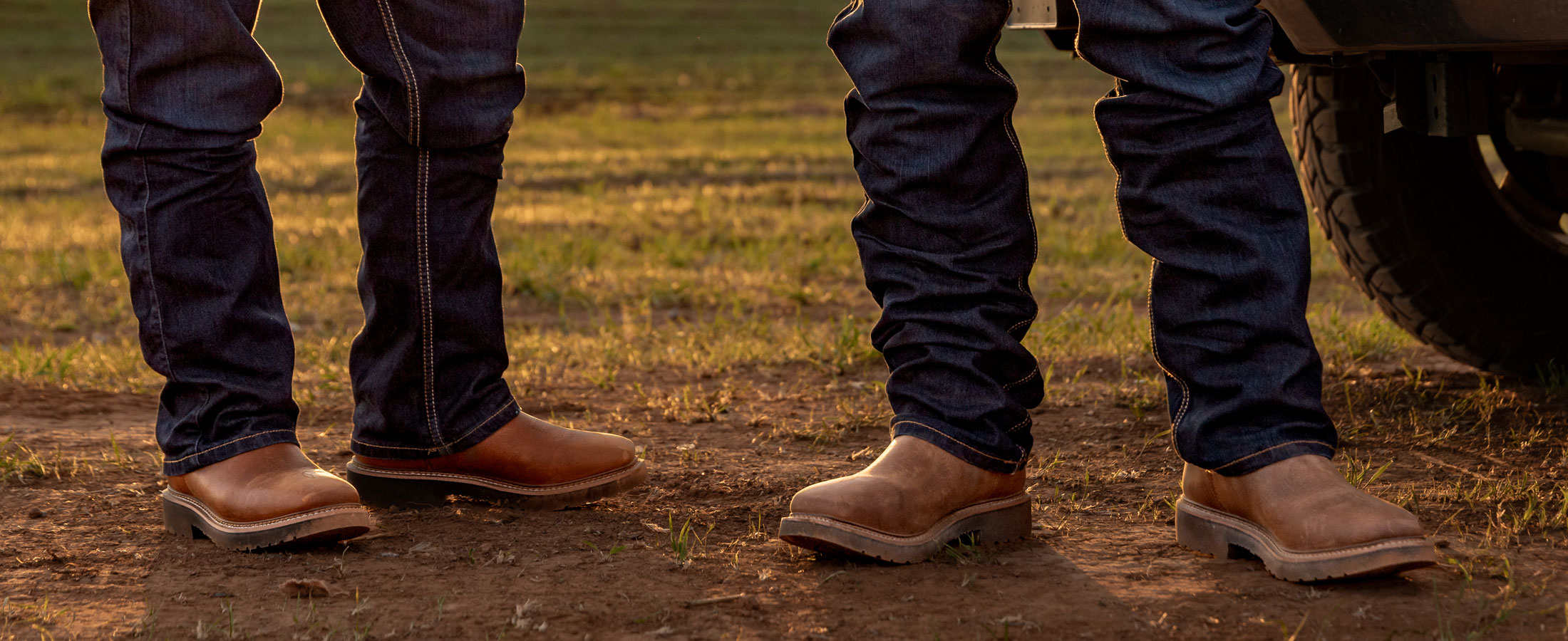 Close-up of two men wearing brown Homeland work boots and jeans, standing on dirt near a truck at sunset.